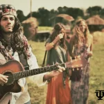 A man dressed in 60's fashion hippie style plays an acoustic guitar outdoors, wearing a colorful headscarf, sunglasses, and layered accessories, with friends in similar attire in the background at a field.