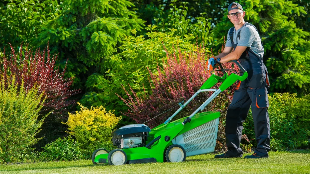 A close-up of a red riding lawn mower cutting grass near a curb, with clippings flying.