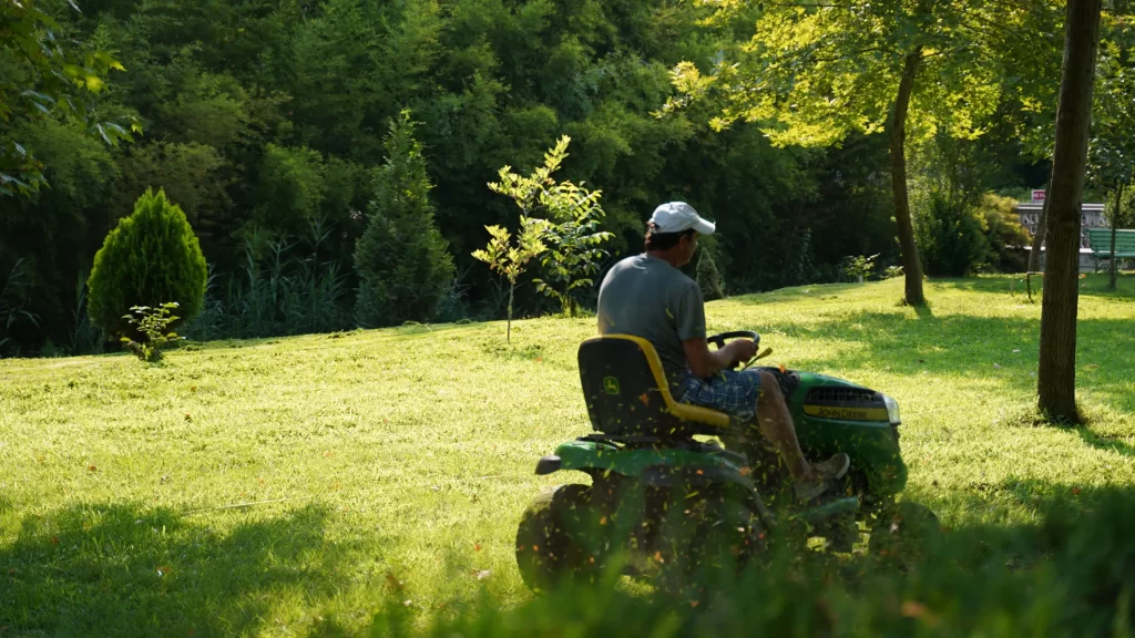 A man mowing the lawn on a John Deere lawn tractor in a well-kept yard with lush greenery.