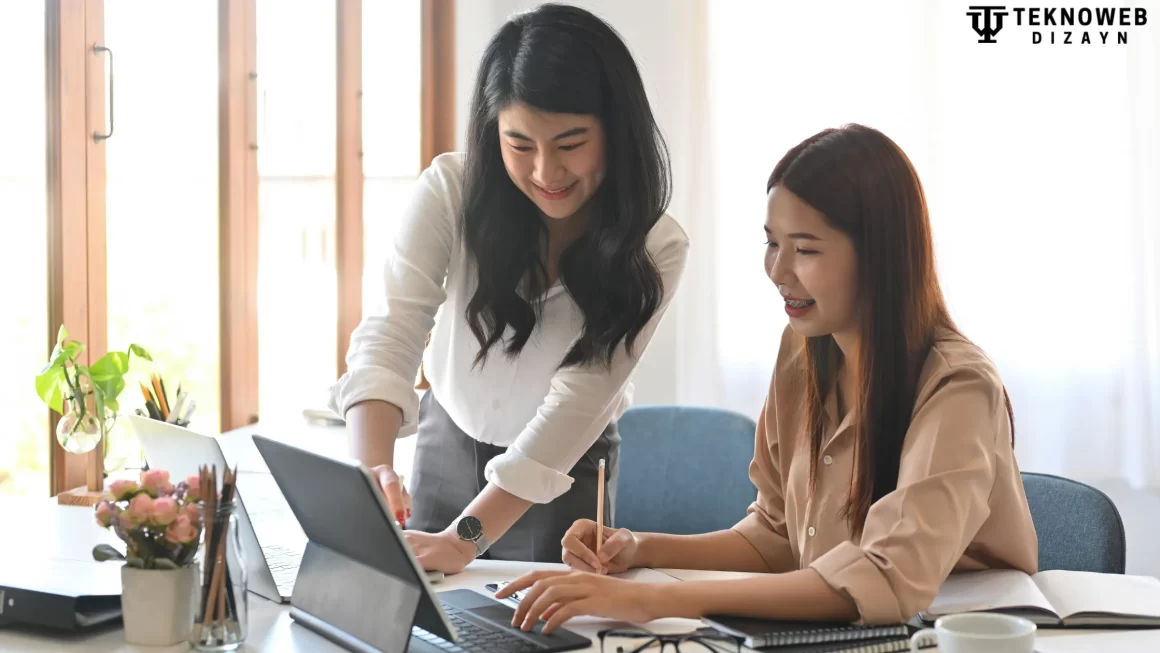 Two female consultants collaborating on a laptop at a cozy workspace with notebooks and office decor.