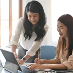 Two female consultants collaborating on a laptop at a cozy workspace with notebooks and office decor.