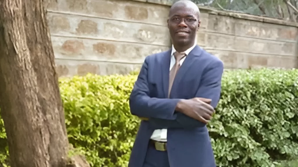 Steven Maranga Nyambega standing confidently outdoors in a suit, arms crossed, in front of a green hedge and brick wall